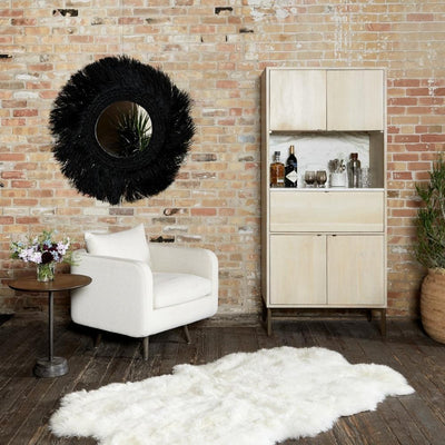 room scene with light wood bar cabinet, white arm chair, round side table, white fluffy rug. Black round mirror on brick wall.