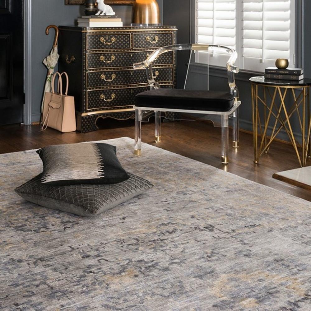 two pillows on area rug with acrylic chair, gold side table, and dark chest.