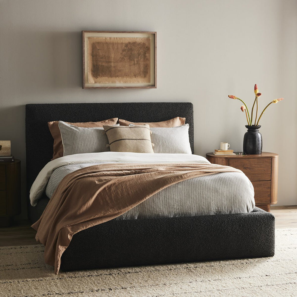 dark fabric bed with blankets and pillows next to wood nightstand