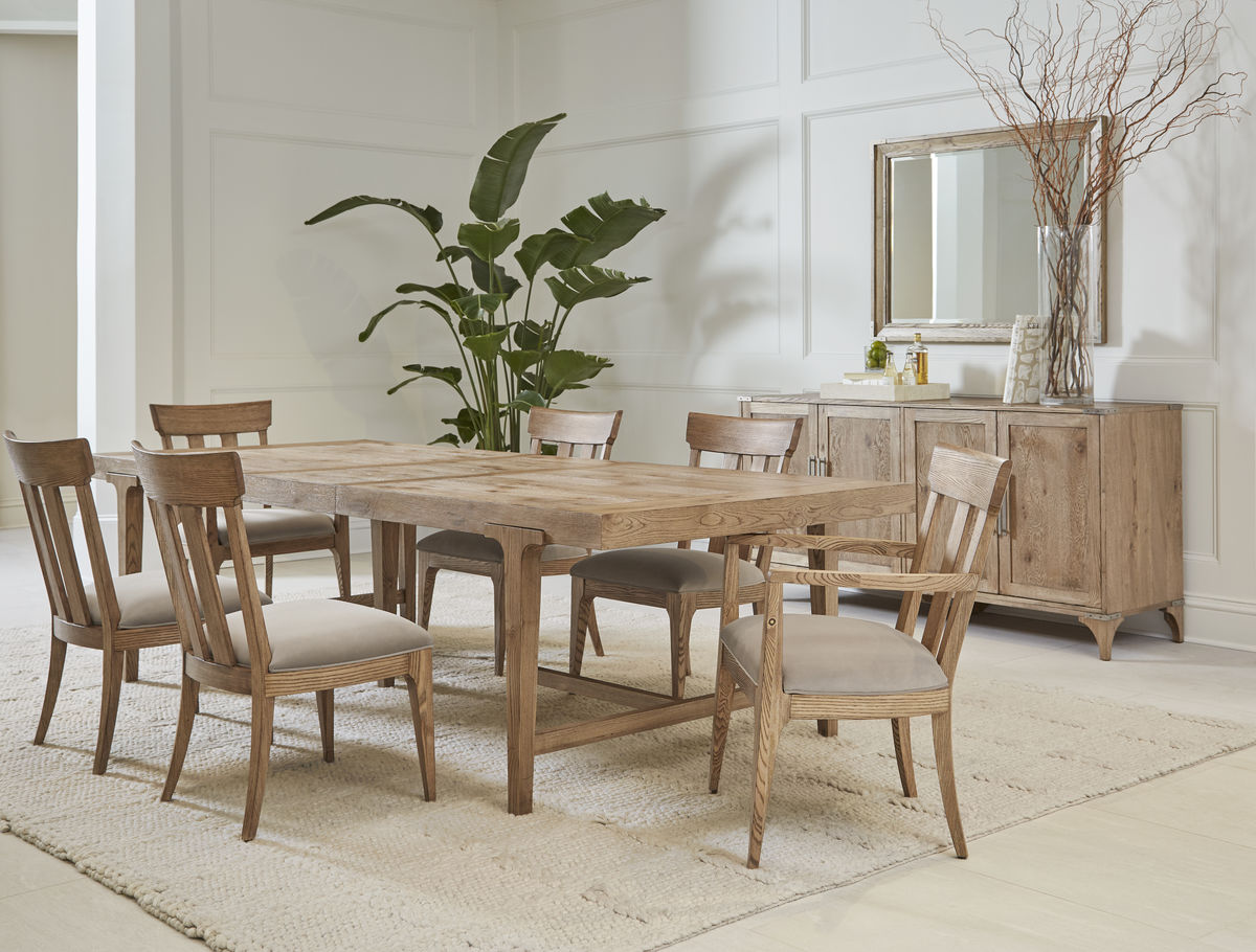 A light wood, rectangular dining table is surrounded by matching dining chairs with beige fabric seats. A matching light wood buffet sits against the side wall under a large square mirror.