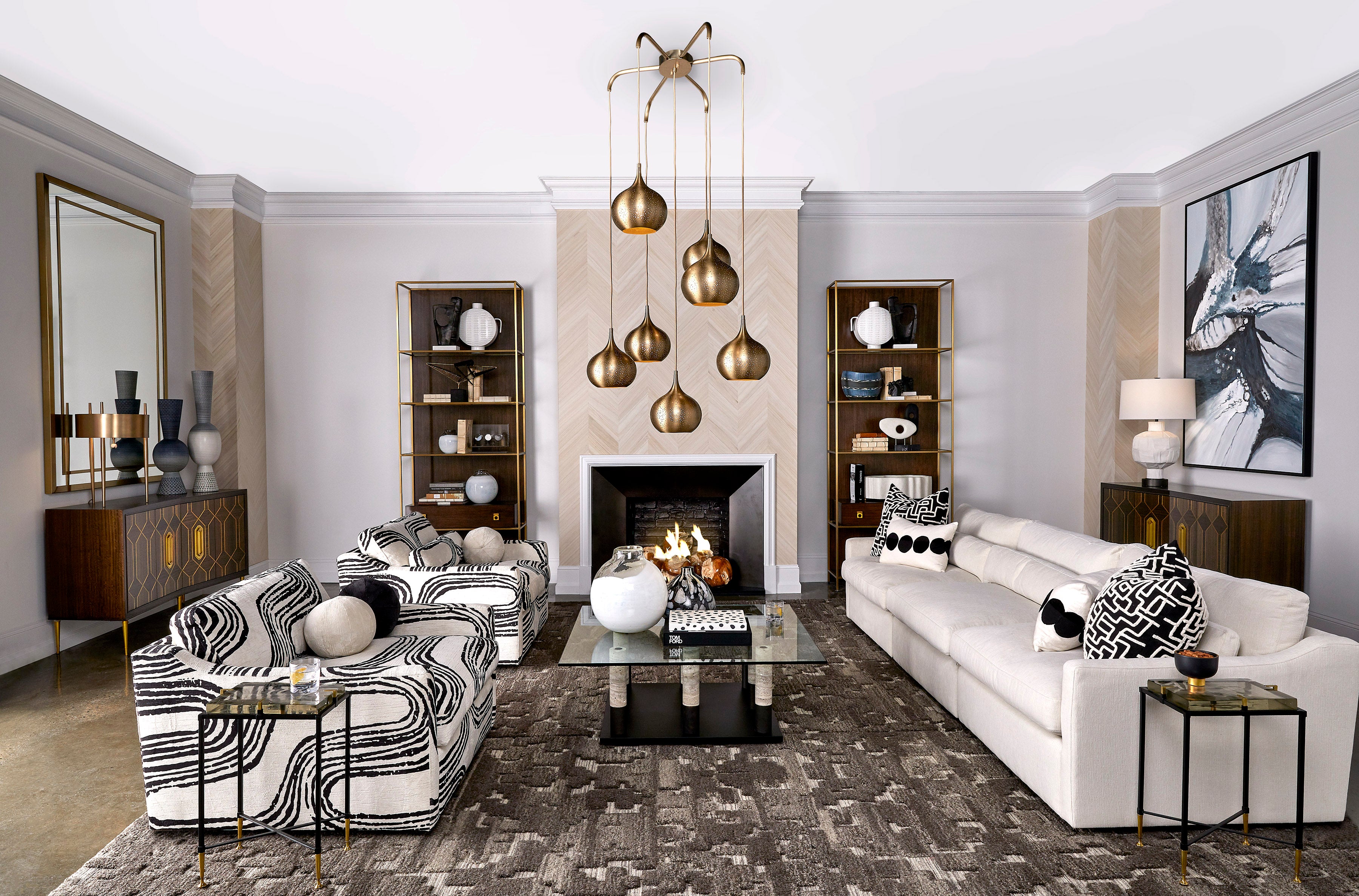 A modern living room with a white sofa across from two black and white geometric print arm chairs and a glass coffee table in between. A large brass chandelier hands from the ceiling while various lamps and decorative pieces litter the room.
