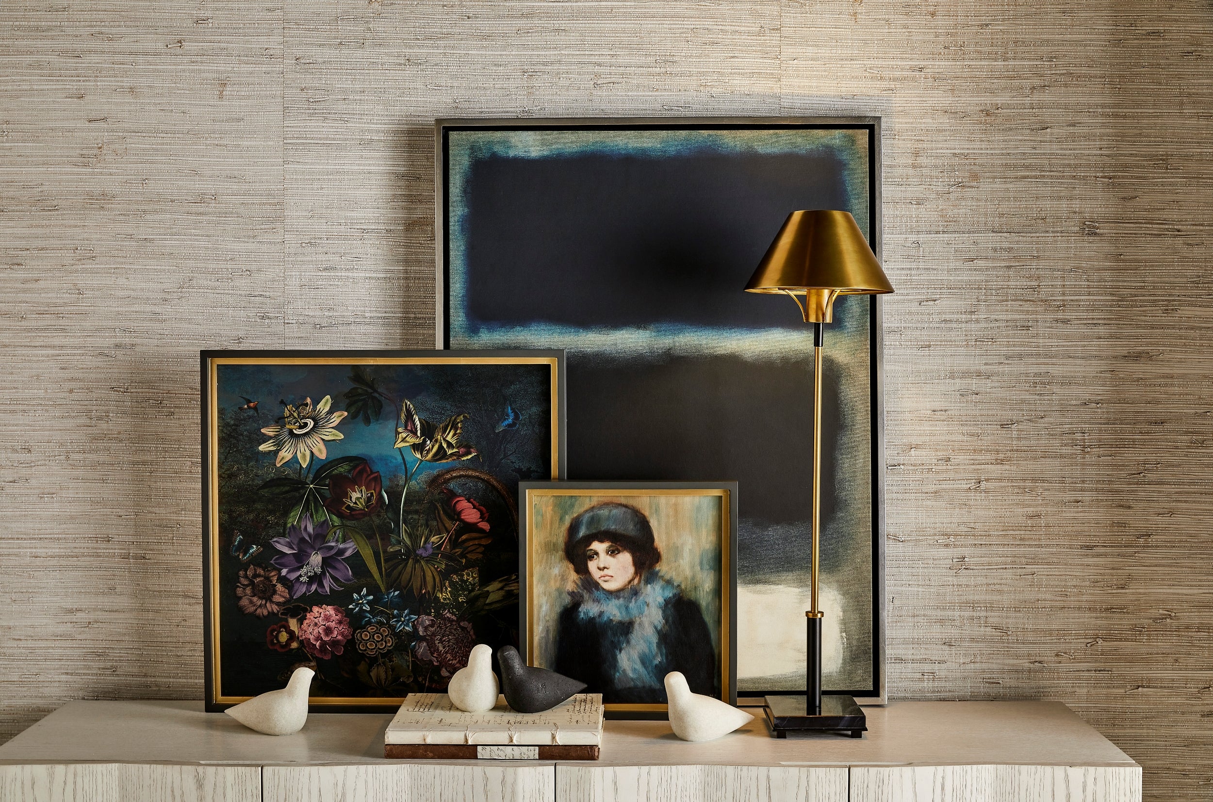 Artwork leaning against a wall atop a credenza cabinet with carved stone bird decor, some books, and a lamp.