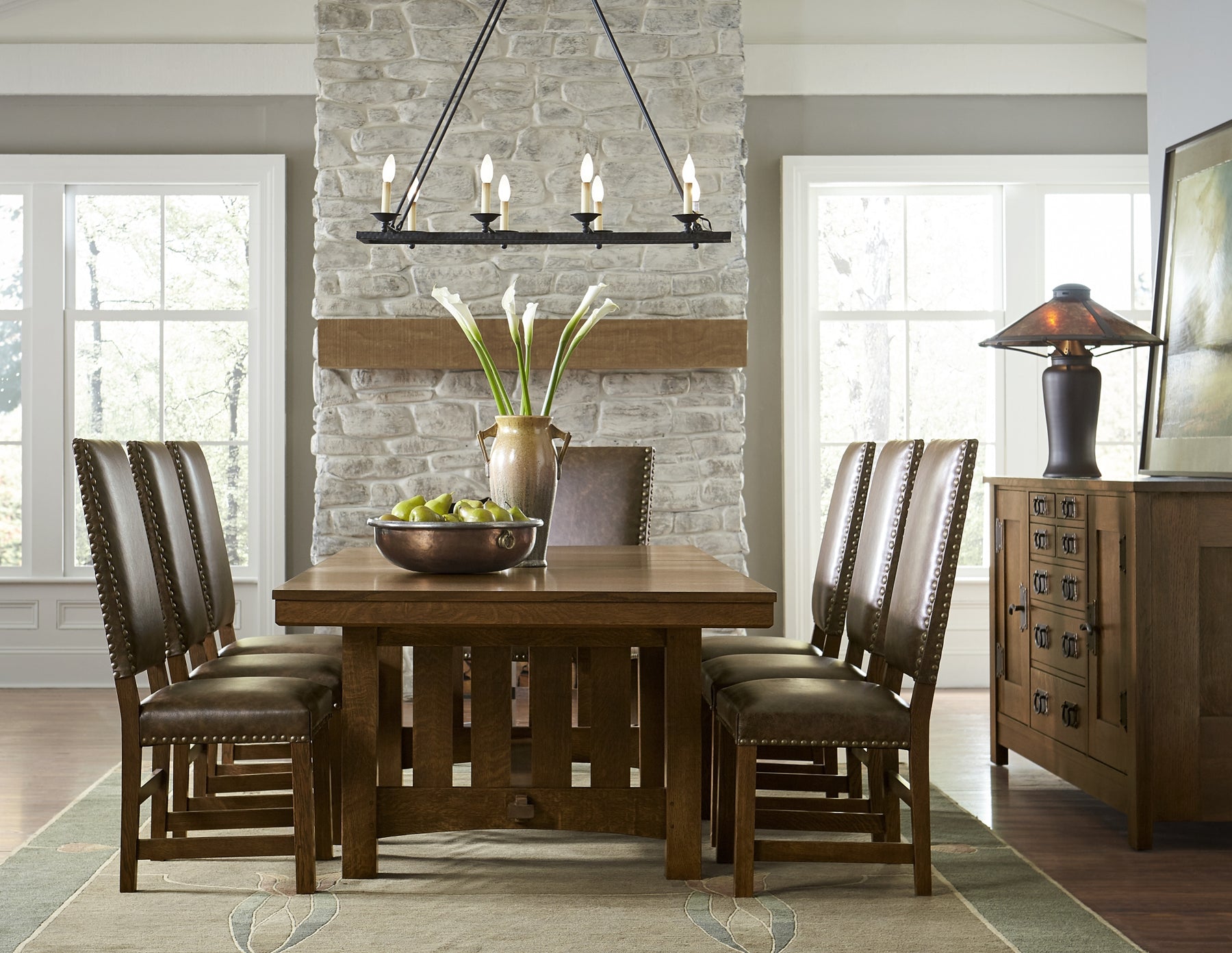 Traditional, rustic dining room featuring a large, rectangular dark wood dining table surrounded by leather upholstered chairs. A metal light fixture hangs from the ceiling and dark wood buffet cabinet sits against the right-hand wall.