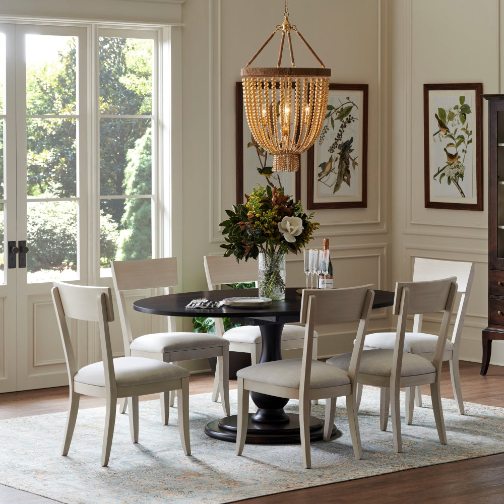 A small dark wood round dining table is surrounded by six white dining chairs. A beaded chandelier hands from the ceiling.