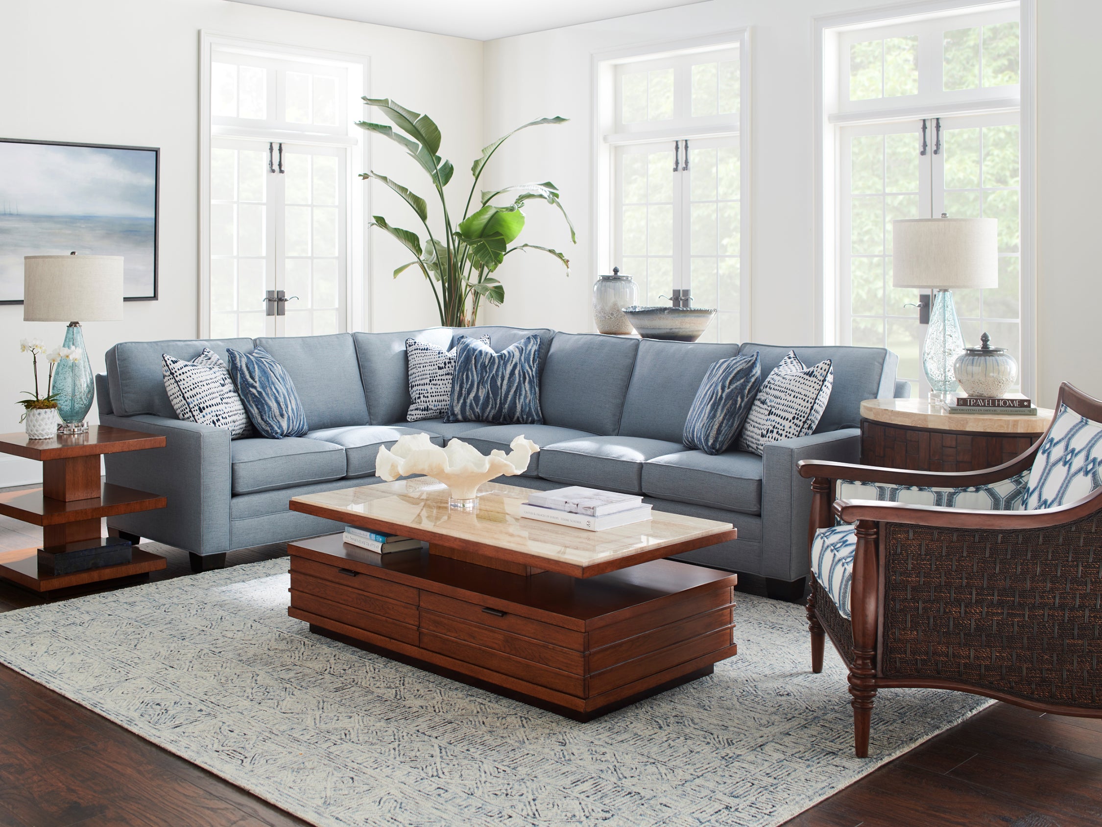 A living room scene from Tommy Bahama Home's Ocean Club collection featuring a large blue fabric sectional sofa and dark wood occasional tables.