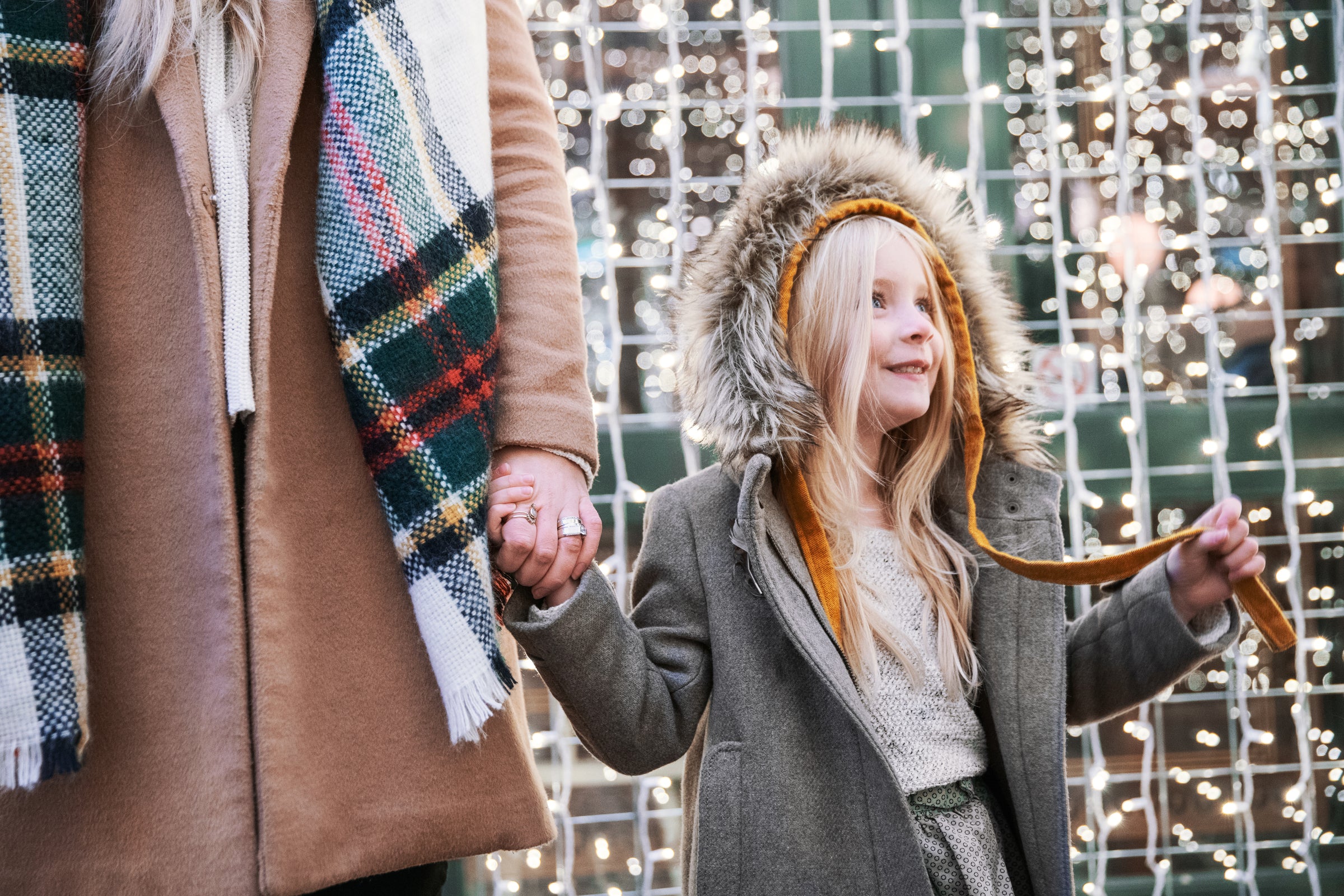 Child in grey coat with fur hood and yellow hat holding hands with adult in beige coat with plaid scarf in front of a wall of string lights