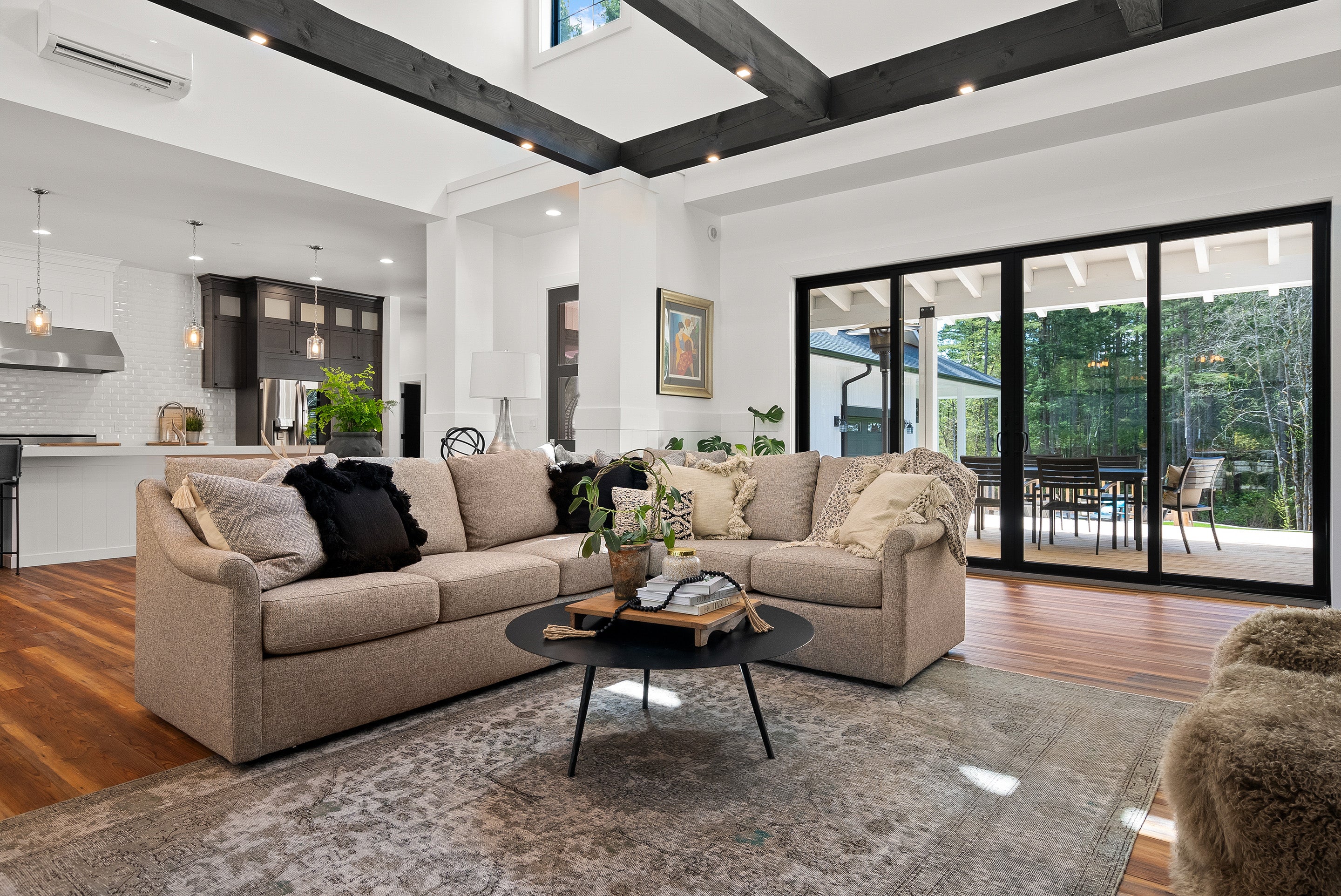 A light brown sectional sofa sits in a large open living room with beige throw pillows. There is a round black coffee table in front of the sofa.