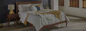A fully made bed with a white comforter and blue and yellow accents on a medium wood bed with a matching nightstand
