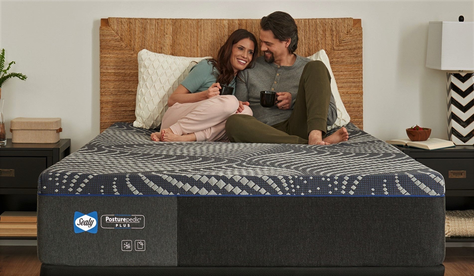 Two people sitting on a Sealy mattress in a bedroom while smiling and holding coffee mugs