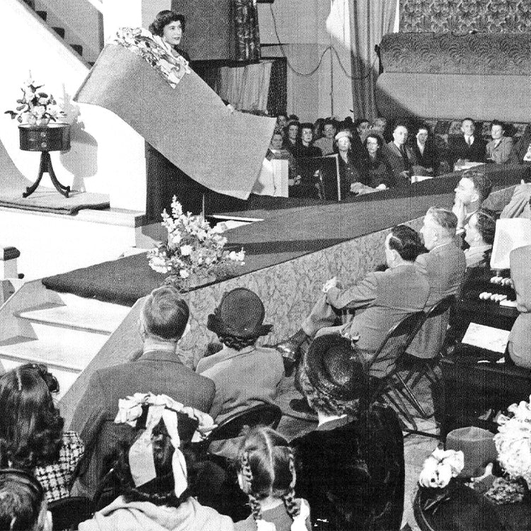 A furniture fashion show circa the 1950's. A woman displays a carpet sample to a crowd of people.